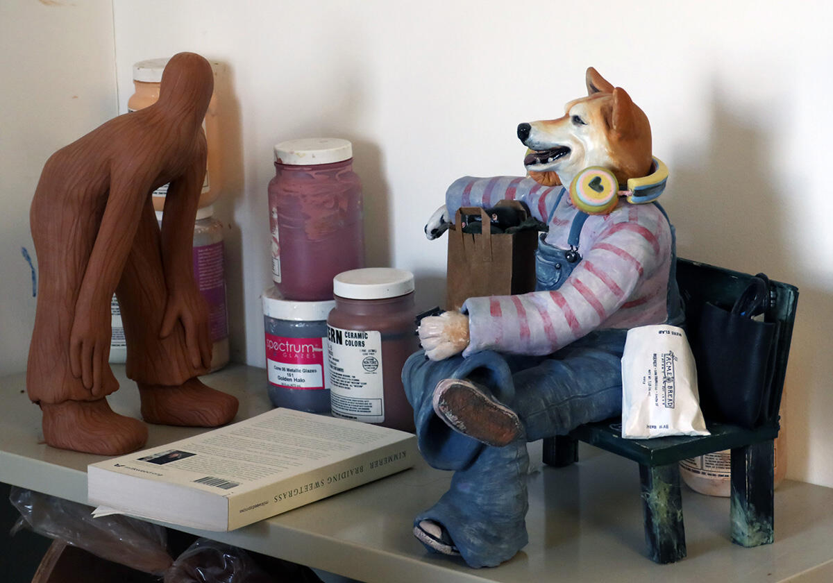 A terracotta figure bends over facing a dog-human hybrid sculpture that is sitting on a park bench with bread and headphones